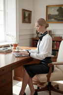 Femme portant une chemise ANI ARPÉ blanche en coton avec col plissé et nœud noir, assise à un bureau en bois.