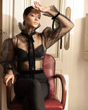 Chemise transparente ANI ARPÉ en plumetis noir avec col montant et ruban noué, portée par une femme assise
